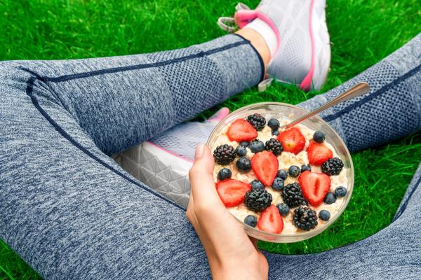 View of woman's legs in gray workout leggings while sitting in the grass holding a bowl of oatmeal and berries